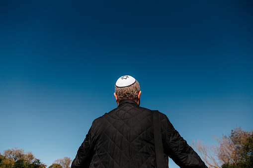 Color image depicting a mid adult Jewish man in his 30s wearing a traditional Jewish skull cap (with star of David design). Rear view low angle image depicting the man looking up at a clear blue sky.