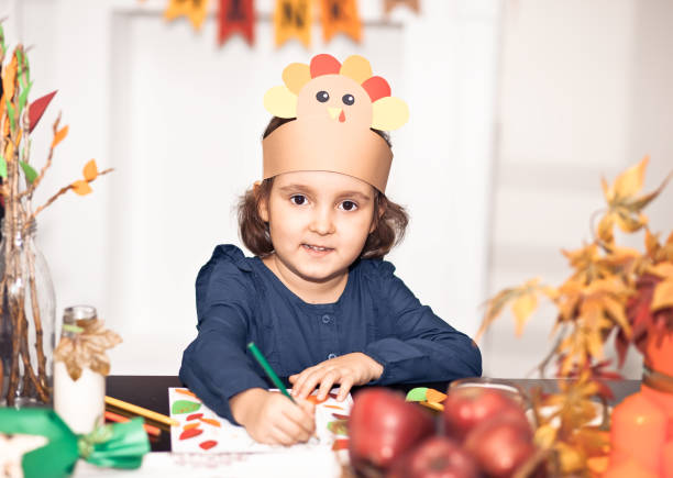 Little cute girl in paper turkey hat writing I am thankful for. Celebrating Thanksgiving day. Diy craft art project. stock photo