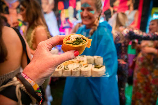 A point of view shot from an unrecognisable caucasian woman dressed in festival clothing, accessories and makeup on an overcast summers day. She is holding a wrap sandwich at a festival themed hen party in a large marquee surrounded by friends.