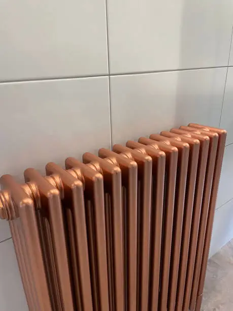 Stock photo showing an old fashioned Victorian-style, copper radiator in front of tiled wall of a modern, luxury bathroom. The radiator is pictured free standing on a stone tiled floor.