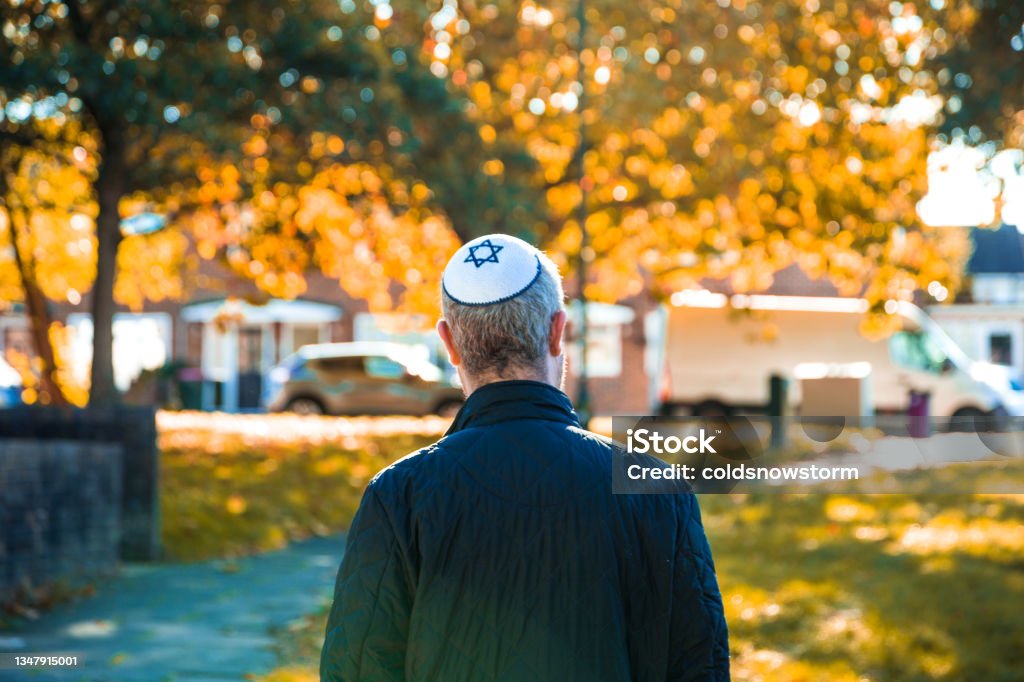 Jewish man wearing skull cap walking on residential street Color image depicting a mid adult Jewish man in his 30s wearing a traditional Jewish skull cap (with star of David design). Rear view of the man as he walks on a residential city street, with houses defocused in the background. Hasidism Stock Photo