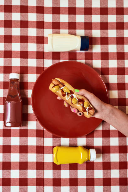 Top view of female hand holding freshly made hot dog with mustard and ketchup over red gingham table cloth