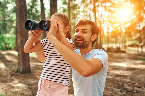 Young dad teaches his little daughter to look through binoculars in a pine forest. Camping, recreation, hiking.
