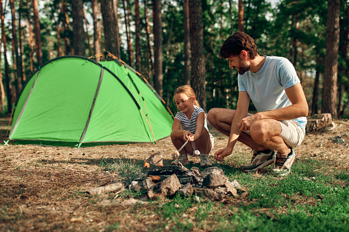 A young father and his daughter sit by a campfire near a tent and grill marshmallows during the weekend in a pine forest. Camping, recreation, hiking.