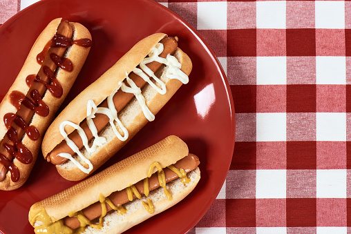 Flatlay of tasty hot dogs with mustard and ketchup on a plate over red checkered plaid napkin. Junk food concept. Horizontal shot