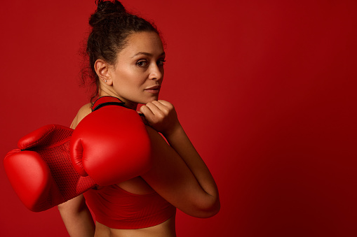 Attractive wavy haired pretty female athlete, woman boxer fighter poses against colored background with red boxing gloves looking confidently at camera through her shoulder. Martial art concept