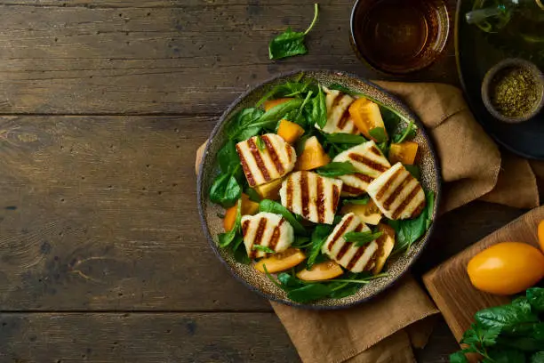Healthy vegetarian summer salad with orange tomatoes, fried circassian halloumi cheese, fresh spinach, and spices on dark brown wooden background. Horizontal, copy space, above