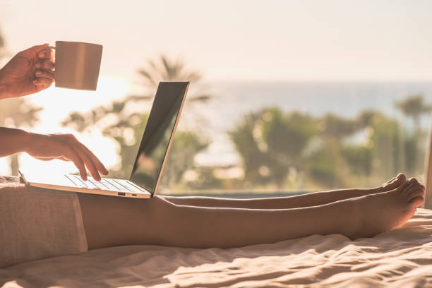 donna freelance che lavora con il computer portatile tenendo una tazza di caffè seduta sul letto in camera da letto vicino alla finestra panoramica con vista sulla spiaggia di mare di palme all'alba - city of sunrise immagine foto e immagini stock