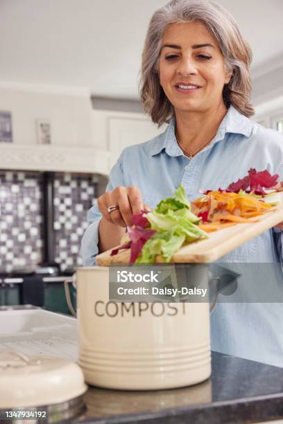 Mature Woman In Kitchen Making Compost Scraping Vegetable Leftovers Into Bin Stock Photo - Download Image Now