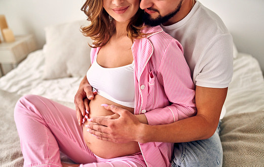 Young pregnant woman sitting on the bed in the bedroom with her husband, who hugs her tummy. Maternity concept.