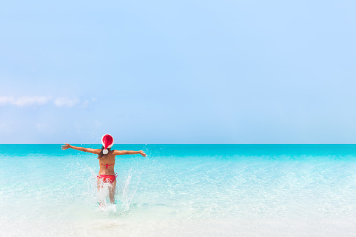 Christmas Caribbean beach tropical getaway Santa Claus woman. Happy Asian bikini girl running winning in success. Open arms freedom carefree. Travel vacation holidays under the tropical sun.