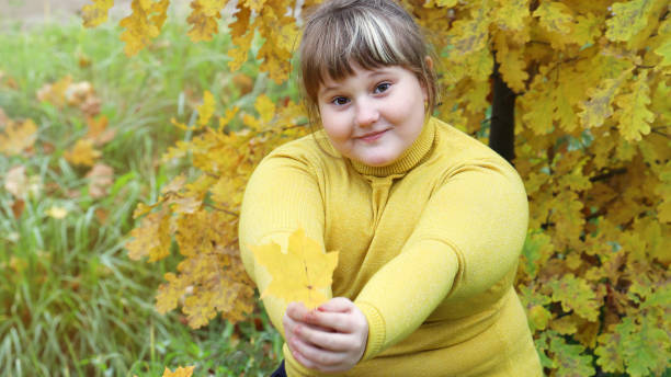 obese smiling girl in yellow clothes give maple leaf forward, on outdoors autumn in yellow foliage - child obesity imagens e fotografias de stock