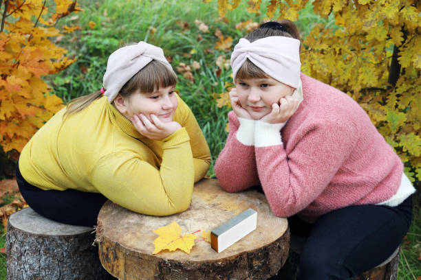 two plump smiling girls sit on stumps outdoors in autumn and listen to music - child obesity imagens e fotografias de stock