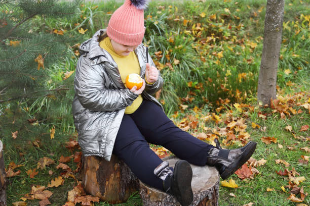 plump smiling girl sits on stump outdoors in autumn day and cleans orange - child obesity imagens e fotografias de stock
