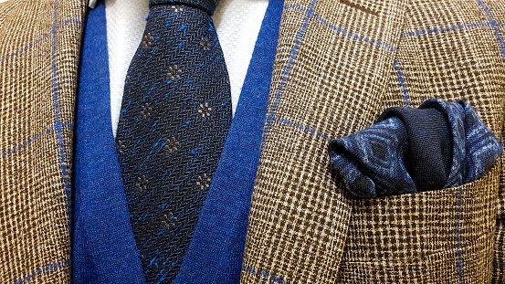 Brown suit jacket with blue squares over a light blue buttoned shirt and a sweater vest together with a matching blue cotton pocket square and a dark blue floral tie