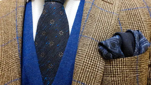 Photo of Brown suit jacket with blue squares over a light blue buttoned shirt and a sweater vest together with a matching blue cotton pocket square and a dark blue floral tie