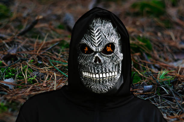 nahaufnahme porträt des sensenmanns. mann in totenmaske mit feuerflamme in den augen auf dunklem wilden naturhintergrund. faschingskostüm, gruselige zähne. halloween feiertagskonzept. dunkler horror - inferno fire flame skull stock-fotos und bilder