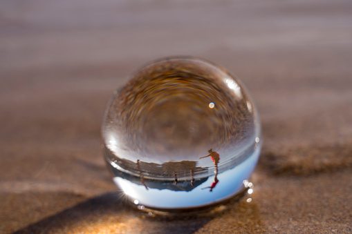 Crystal ball resting in wet sandy beach, red sand, reflecting a person and sky