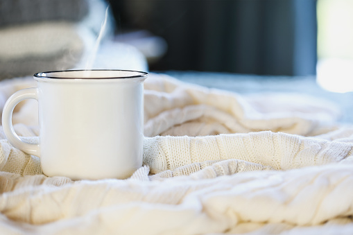 Hot steaming cup of coffee sitting on top a soft white knit blanket on a bed with stack of covers in background. Selective focus with extreme blurred foreground and background.