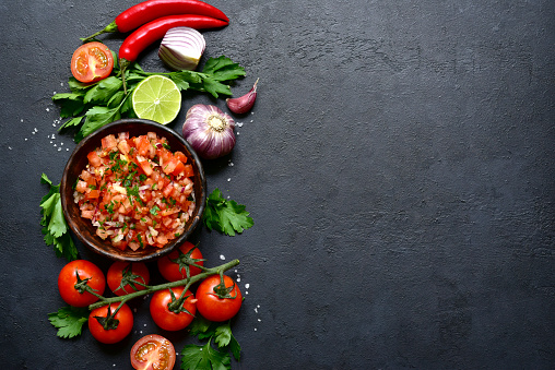 Tomato salsa (salsa roja) - traditional mexican sauce  with ingredients for making on a dark  slate,stone or concrete background.Top view with copy space.