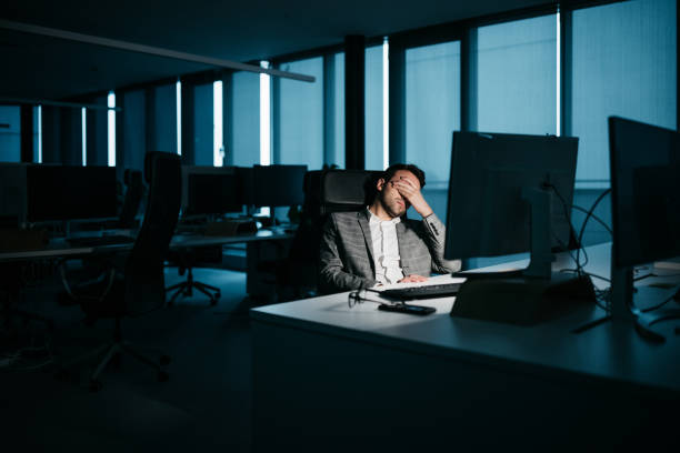 Young businessman tired at night A smartly-dressed businessman is resting in his office in front of his computer. He is tired and wants to go home. His face is not recognizable. Low-key lighting. Horizontal night-time indoor photo. mental burnout photos stock pictures, royalty-free photos & images