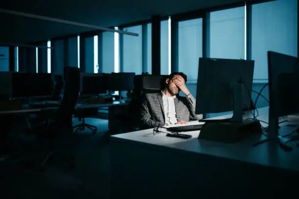 A smartly-dressed businessman is resting in his office in front of his computer. He is tired and wants to go home. His face is not recognizable. Low-key lighting. Horizontal night-time indoor photo.