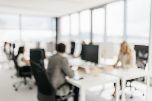 A blurred photo of businnes colleagues working on their computers. They are sitting behind a large desk in a bright office with large windows. Their faces are not visible. Horizontal daylight indoor photo with copy space.