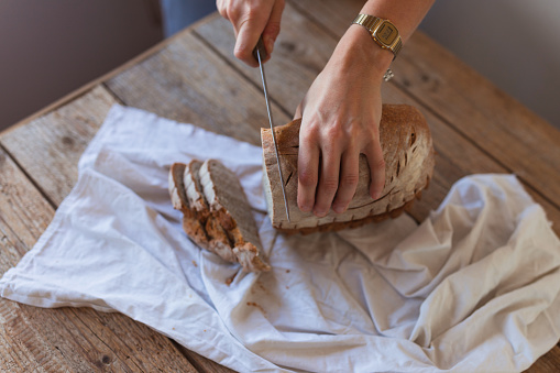 High angle view of an anonymous female baker with apron putting slices of cheese on bread with hummus while standing at kitchen desk with a wooden cutting board and a big knife.