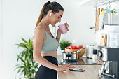 Sporty woman listening to music with her smartphone whiledrinking coffee in the kitchen at home.
