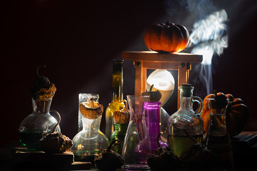 Photo of halloween symbols such as pumpkin, poison bottle, rotten pumpkins and hourglass in dark. Light beam is coming from right side of frame. No people are seen in frame. Shot with a full frame mirrorless camera.