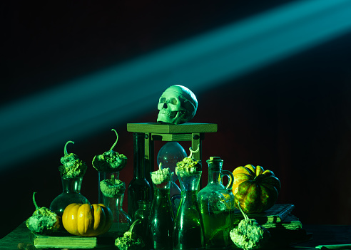 Photo of pumpkin, poison glasses, human skull and hourglass under dramatic light for halloween. No people are seen in frame. A colored light is illuminating objects. Shot with a full frame mirrorless camera.