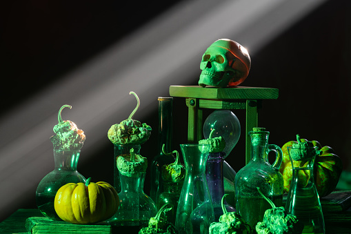 Photo of pumpkin, poison glasses, human skull and hourglass under dramatic light for halloween. No people are seen in frame. A colored light is illuminating objects. Shot with a full frame mirrorless camera.