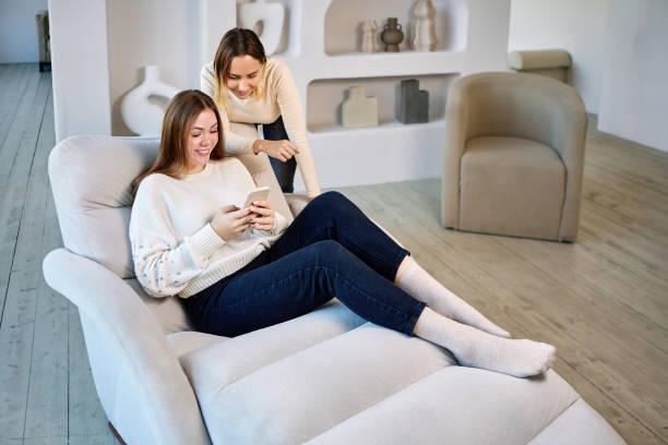 Two women on chaise longue with phone in lounge. Two happy women on sofa are chatting with friends using smartphone in living room. chaise longue woman stock pictures, royalty-free photos & images