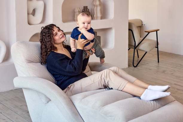 Couch in living room with woman who holds child. Lounger in lounge with mother who is holding infant in hands during sitting on sofa. chaise longue woman stock pictures, royalty-free photos & images