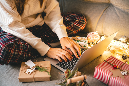 Close up of woman hands with gifts, typing at laptop. Online shopping at Christmas holidays. Cropped female sit on couch with natural eco presents and decor. Merry Christmas packing Concept.