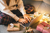 Close up of woman hands with gifts, typing at laptop. Online shopping at Christmas holidays. Cropped female sit on couch with natural eco presents and decor. Merry Christmas packing Concept