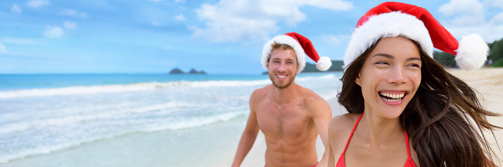 Happy Christmas holiday woman and man honeymoon couple laughing on beach vacation wearing santa claus hats carefree in freedom. Smiling Asian girl people lifestyle.