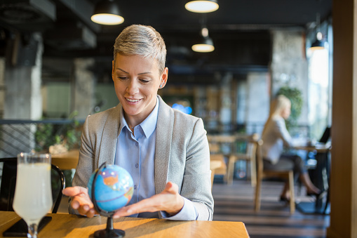 Smiling woman sitting at coffee shop and planning to travel. Smiling businesswoman looking at globe and planning to her new business trip or vacation.