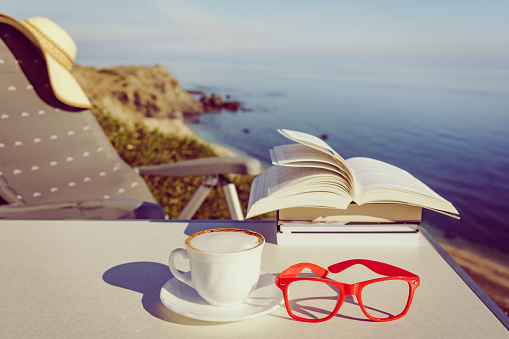 Reading on holidays. Coffee cup and books on table outdoors against blue sea water background. Book pages fluttering in the wind.