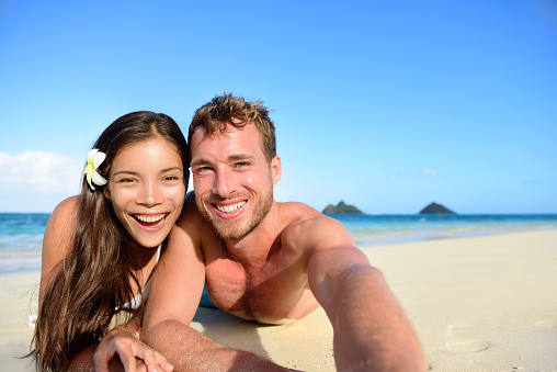 Couple relaxing on beach taking selfie picture with camera smartphone. Young multiracial couple on getaway vacation in Hawaii lying down looking at camera. Candid closeup angle looking real. Lanikai beach