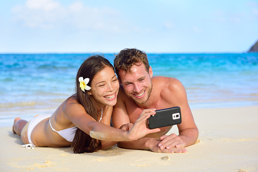 Beach holiday couple taking selfie with smartphone lying down relaxing and having fun holding smart phone camera. Young beautiful multicultural Asian Caucasian couple having fun on summer beach.