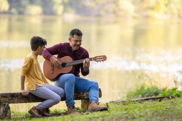 padre e hijo tocando la guitarra frente al lago - plucking an instrument fotografías e imágenes de stock