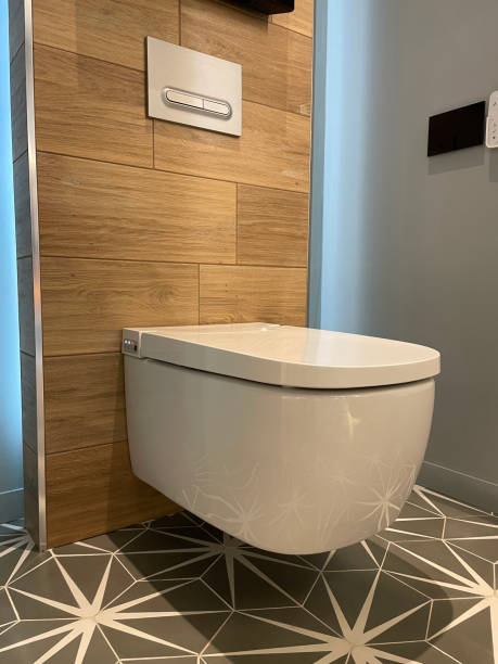 Stock photo showing a modern, white square-shaped wall hung toilet pan, with soft close toilet seat lid, hanging on wooden clad washroom wall. The floor is covered with hexagonal tiles that tessellate to make a large black and white star pattern. imagen de una suite de baño moderna, blanca y de lujo, una sartén de inodoro colgada de pared de forma cuadrada contemporánea con tapa de asiento de inodoro cerrada suave que cuelga de la pared del baño revestida de madera, placa de pared de doble desc