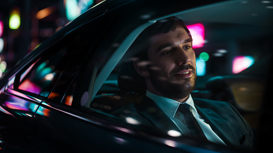 Handsome Businessman in a Suit Commuting from Office in a Backseat of His Car at Night. Entrepreneur Passenger Traveling in a Transfer Taxi in Urban City Street with Working Neon Signs.