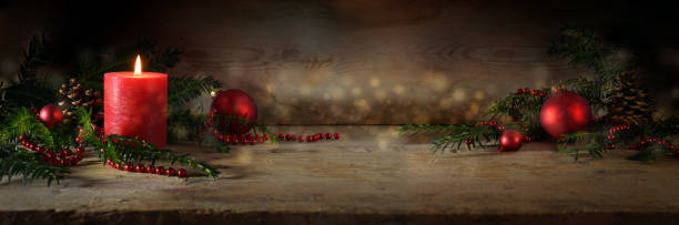 Atmospheric Advent and Christmas decoration with a lit red candle, balls and evergreen yew branches on dark rustic wooden planks, wide panoramic format, copy space stock photo