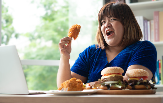 Hungry overweight woman holding Fried Chicken, hamburger on a wooden plate and Pizza on table, During work from home, gain weight problem. Concept of binge eating disorder (BED).