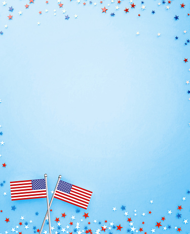 Star shaped confetti and tiny American flag pair on blue background. Vertical composition with copy space. Directly above.