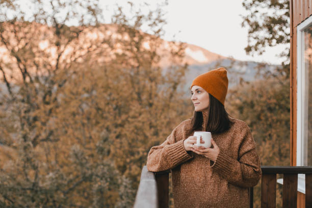 giovane donna in maglia maglione e cappello che beve tè e mangia croissant freschi sull'accogliente balcone di una casa di campagna in legno. - balcony house golf home interior foto e immagini stock