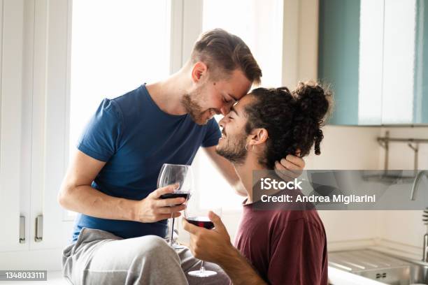 Tenderness Moment Of Smiling Gay Couple Young Men Toasting With Red Wine In The Kitchen Male Couple Having Fun And Drinking Red Wine For Celebration Of Their Engagement Stock Photo - Download Image Now