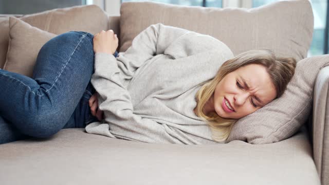 young woman sitting or lying on sofa or couch at home with severe abdominal pain. Female on bed holds her stomach with premenstrual,